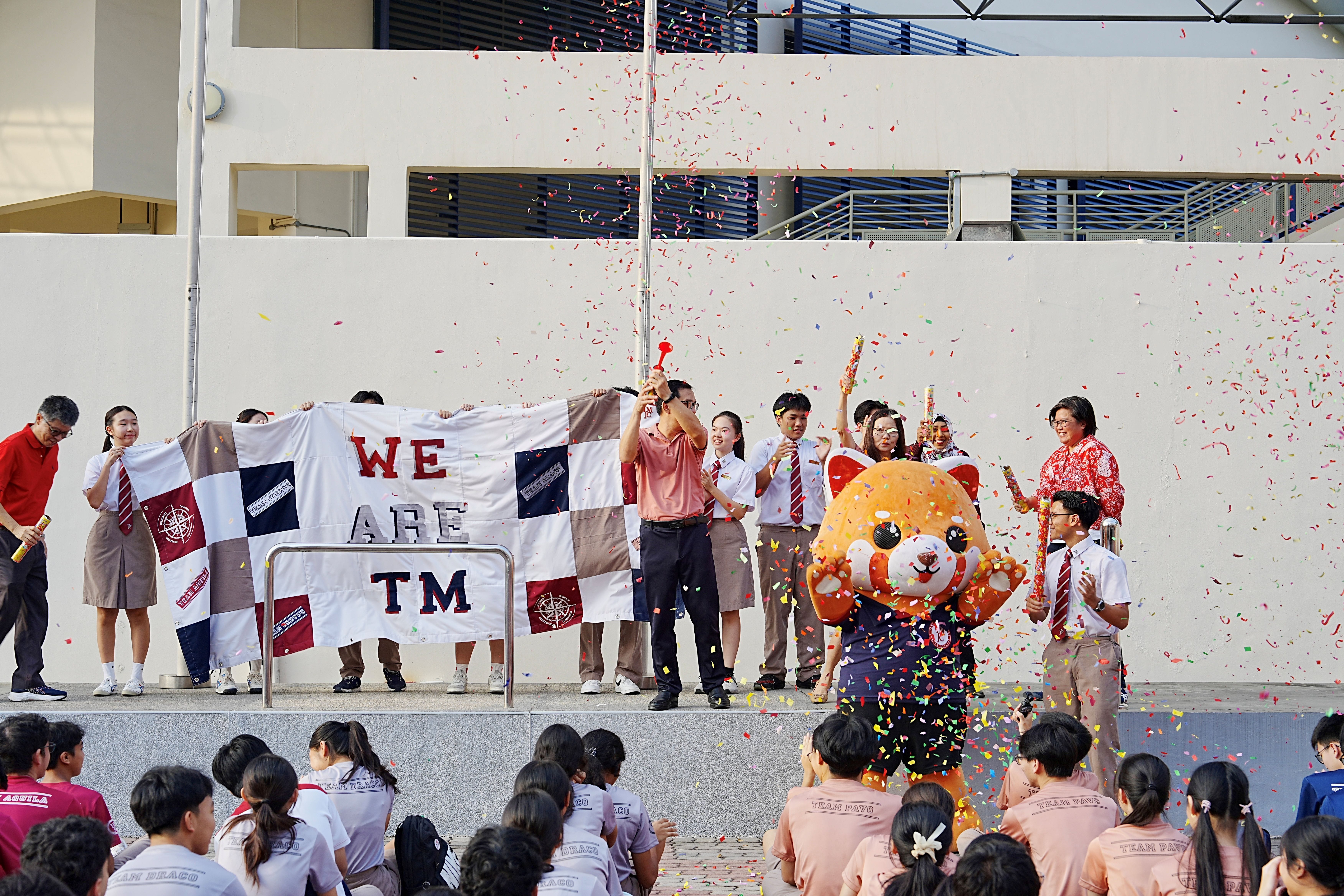 A celebratory outdoor school event with confetti raining down. Students and teachers stand on a raised platform holding a patchwork banner reading "WE ARE TM", alongside a red panda mascot costume character.